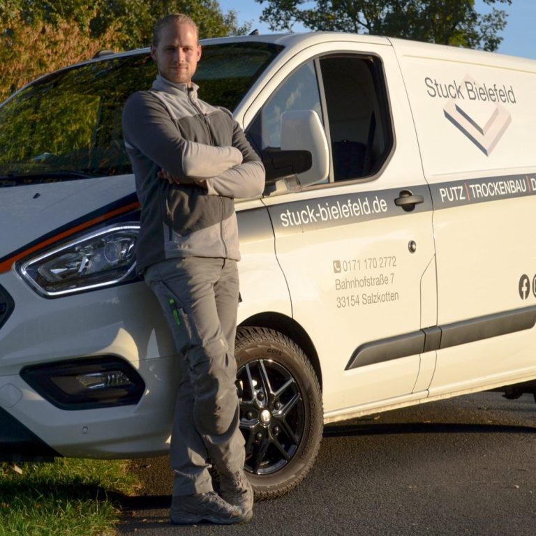 Mann im grauen Arbeitsanzug steht neben einem weißen Transporter mit Firmenlogo.