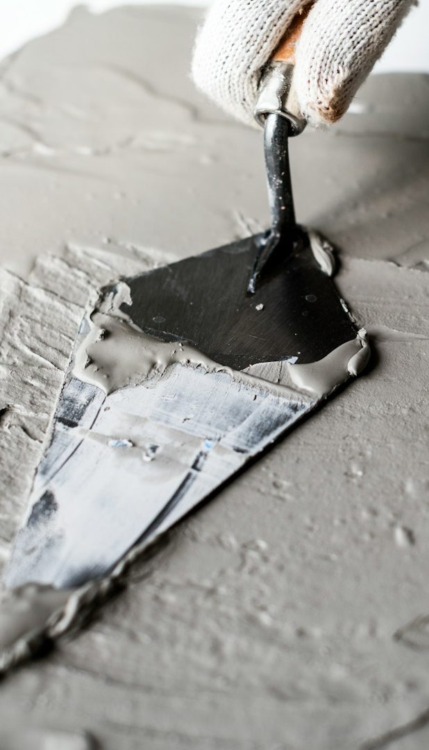 Ein Handwerker trägt schwarzen Mörtel mit einer Kelle auf eine graue Fläche auf.