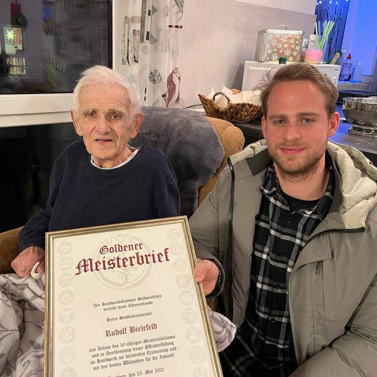 Ein älterer Herr und ein Mann halten einen goldenen Meisterbrief in der Hand, lächelnd zusammen.